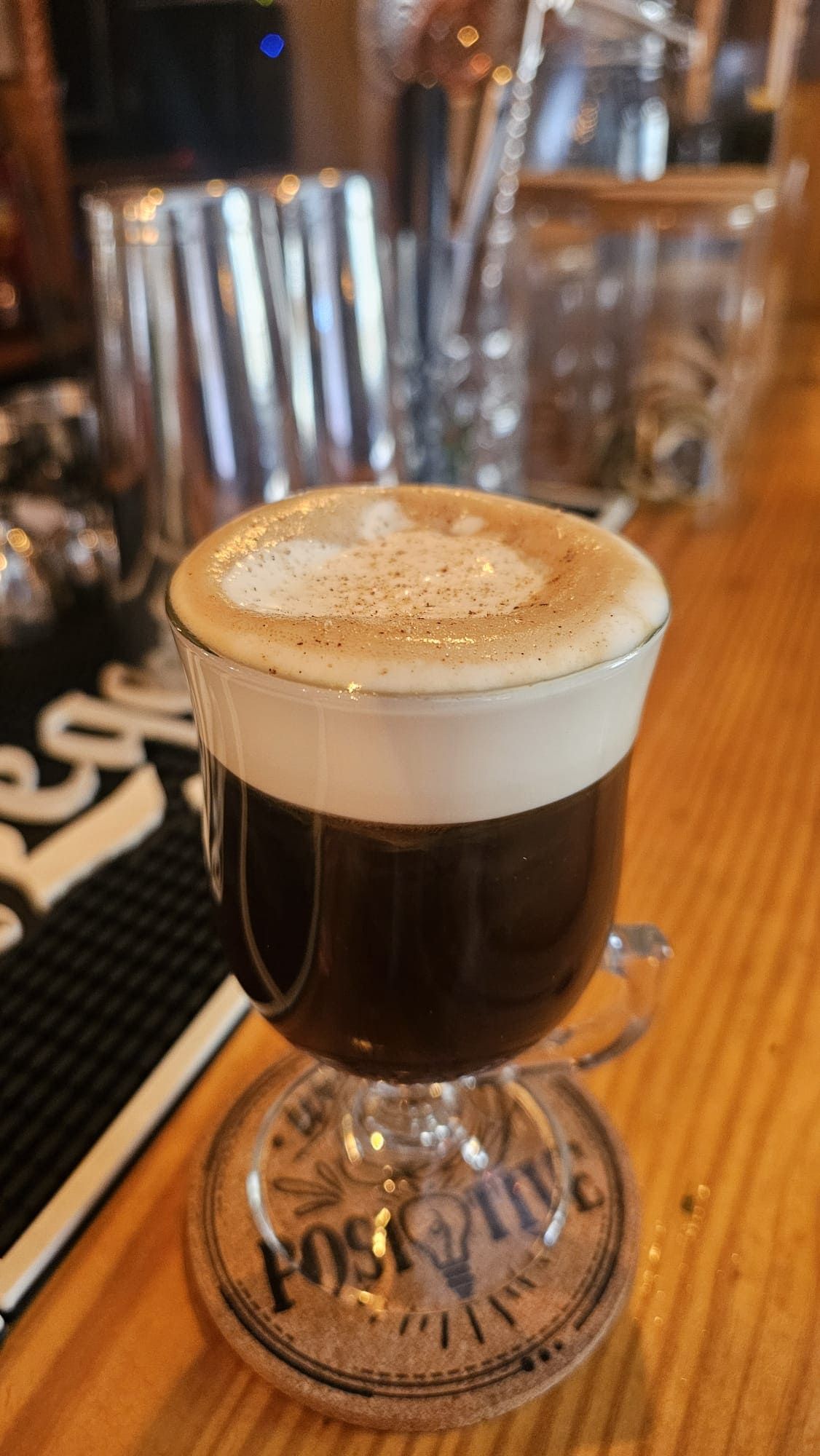 Un vaso de café irlandés con una espesa capa de nata y una pizca de especias, que reposa sobre un posavasos redondo en una barra de madera.