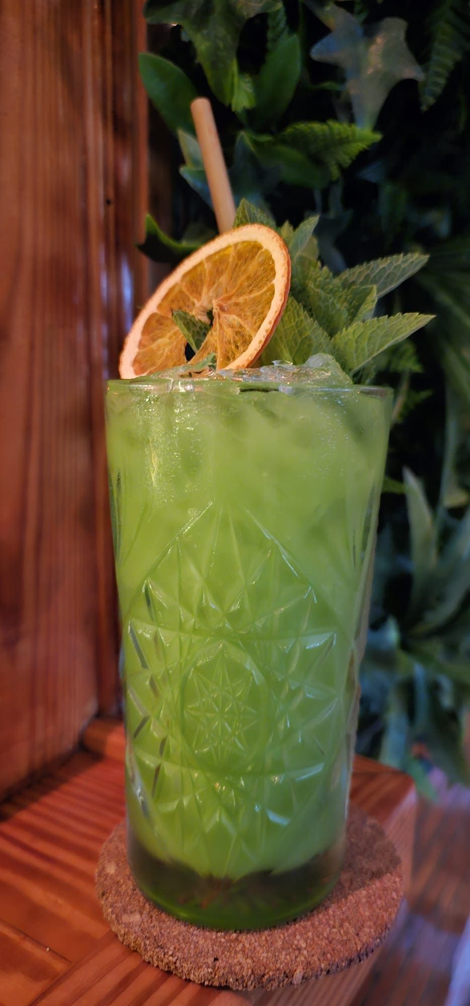 Un cóctel de color verde brillante en un vaso texturizado, adornado con menta fresca y una rodaja de naranja deshidratada, sobre una superficie de madera.
