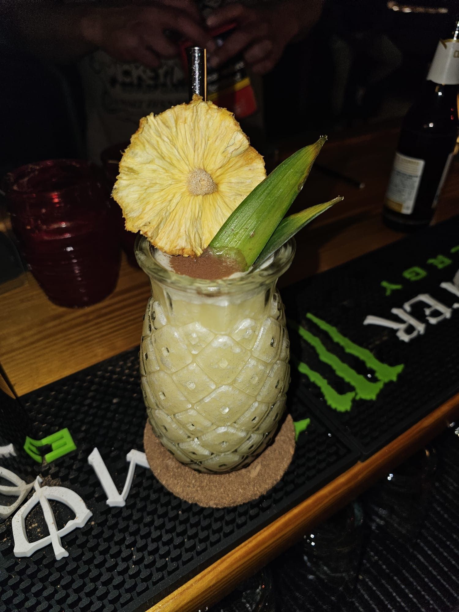 Un vaso con forma de piña lleno de un cóctel de color claro, adornado con una rodaja de piña deshidratada y hojas verdes.