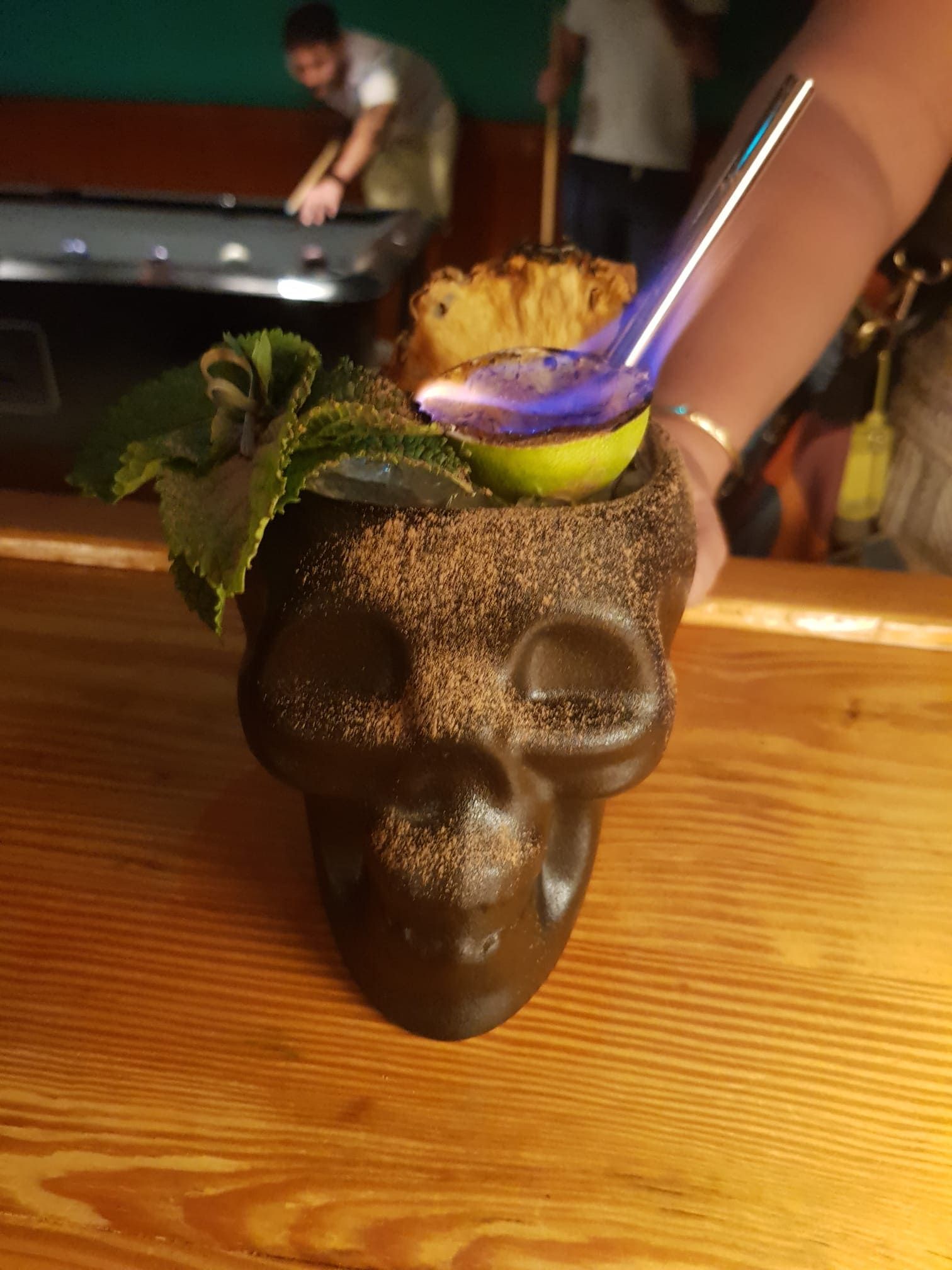 Un cóctel flameado servido en una taza con forma de calavera, adornado con menta y piña, en un bar con una mesa de billar al fondo.