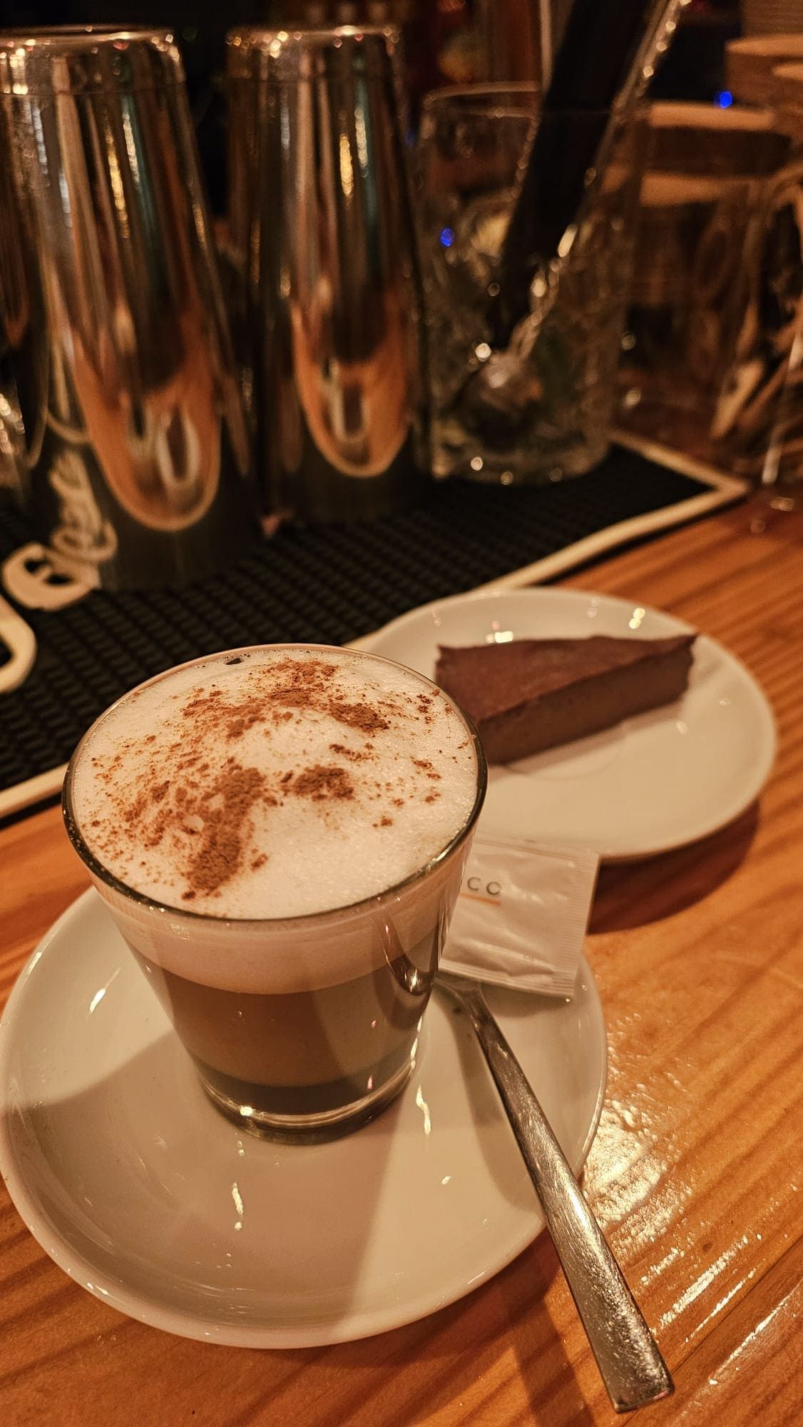 Un vaso de capuchino con cacao en polvo, servido en un platillo con una cuchara y una porción de pastel de chocolate sobre una mesa.