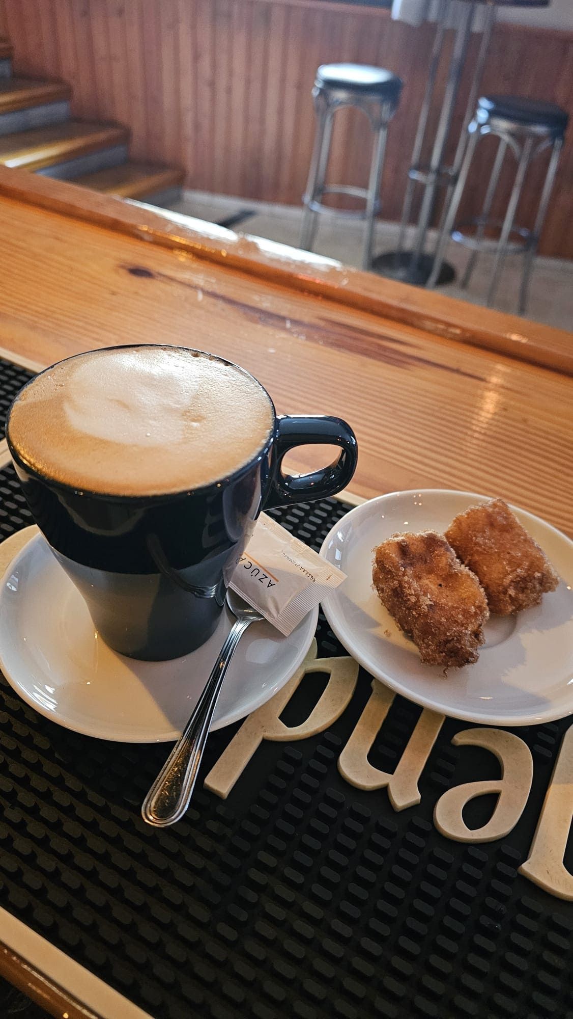Una taza de café oscuro con espuma de leche junto a dos pequeños bocadillos fritos de color marrón dorado sobre un mantel individual en una cafetería.