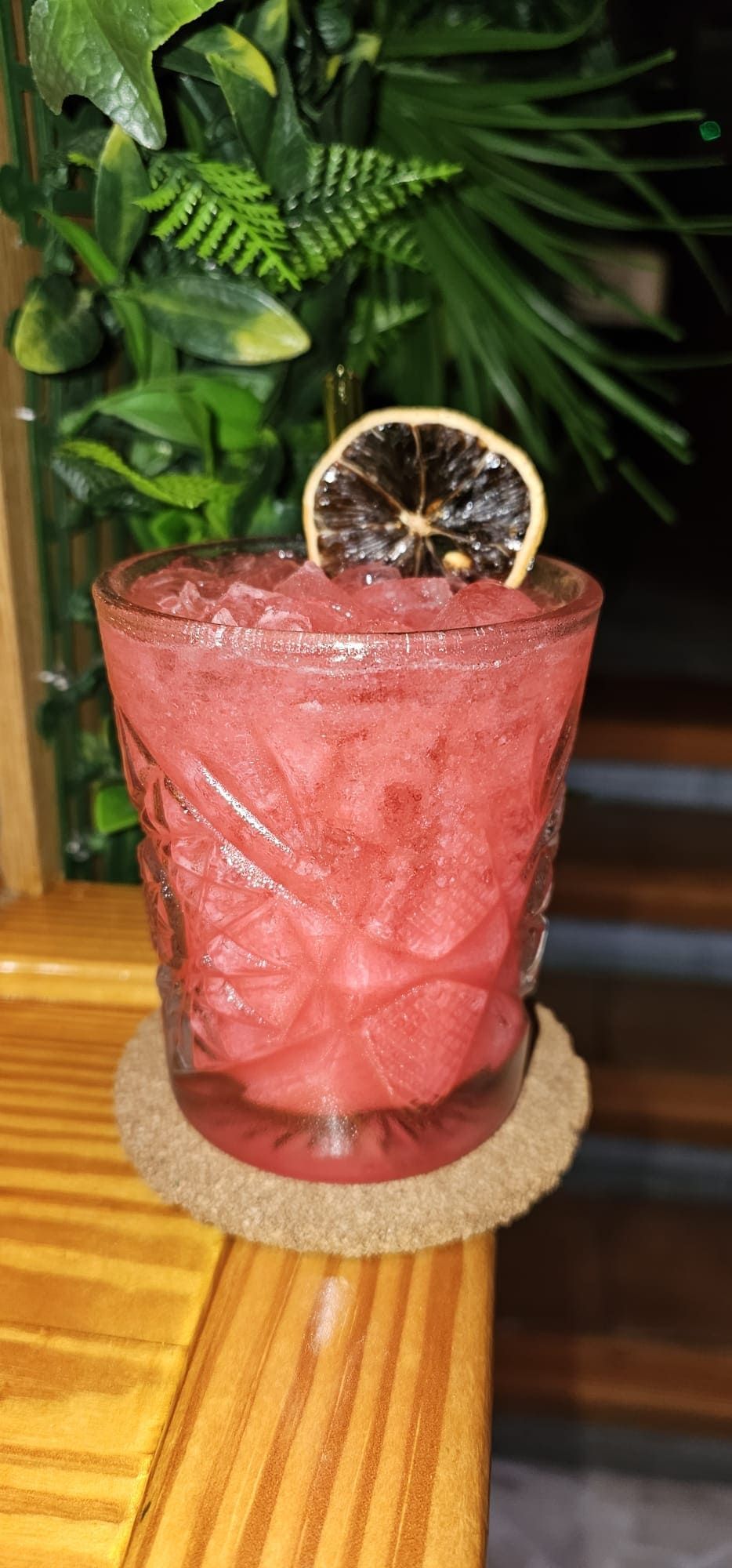 Un cóctel rosa en un vaso texturizado con una rodaja de cítricos deshidratada como guarnición, sobre un posavasos.