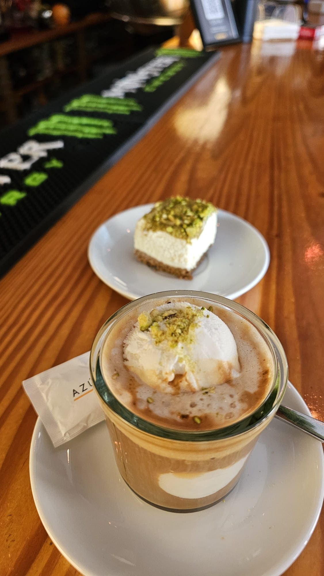 Un café con helado y una tarta de queso con pistachos sobre una barra de madera con una esterilla negra de Monster Energy.