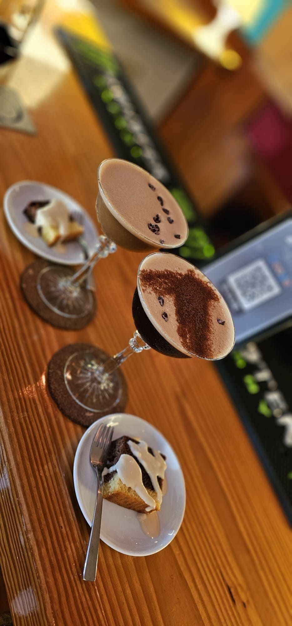 Dos copas de cóctel con espuma y polvo de chocolate sobre una barra de madera, junto a dos platitos con porciones de postre.