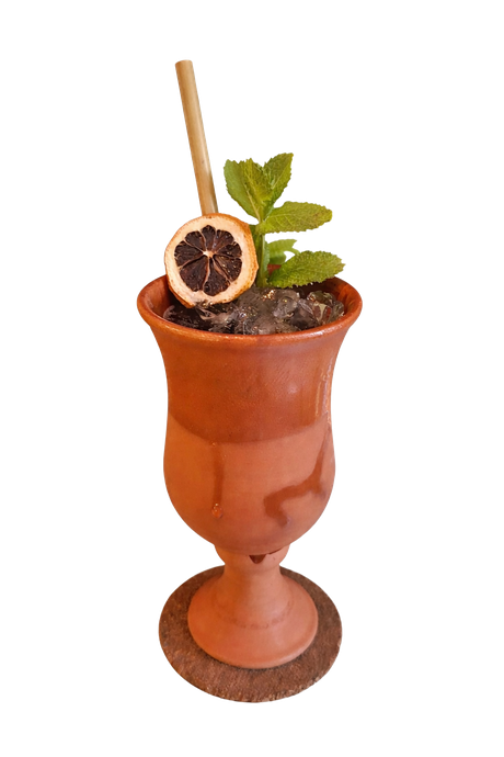 Un cáliz de terracota lleno de hielo, coronado con una rodaja de cítrico deshidratado, una ramita de menta y una pajita sobre un posavasos de corcho.