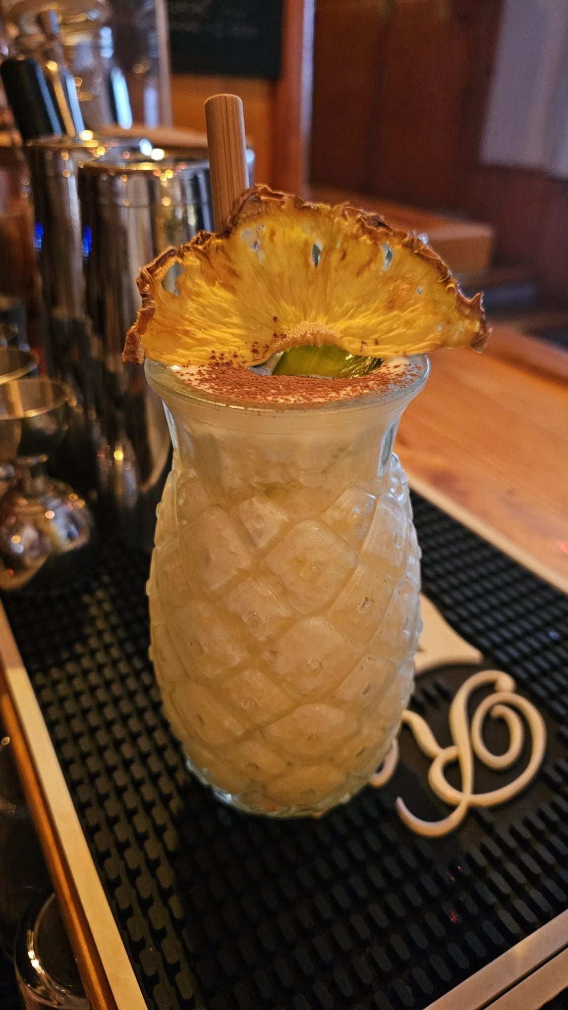 Un cóctel tropical servido en un vaso texturizado con forma de piña, adornado con una rodaja de piña deshidratada y una pajita, sobre una barra.
