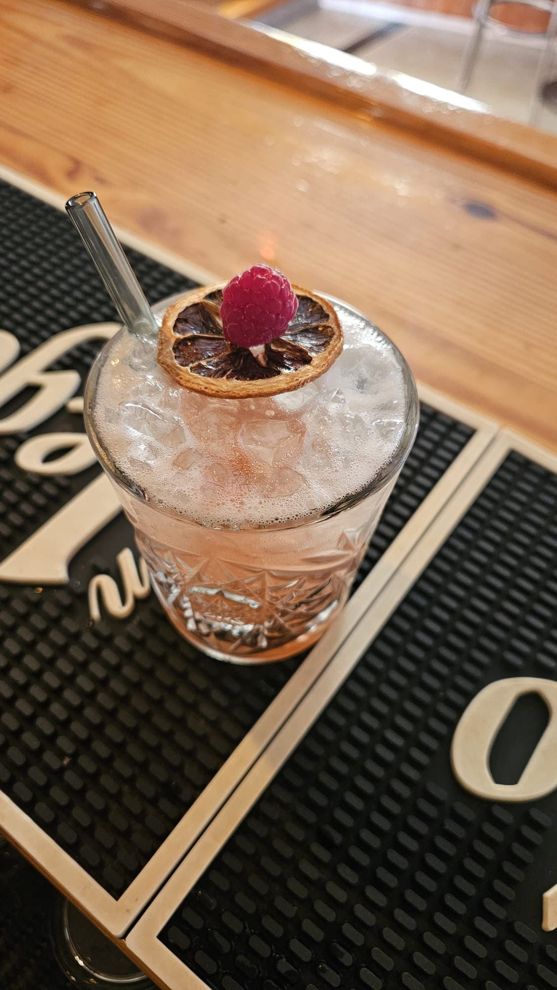Un cóctel artesanal servido en un vaso estampado, adornado con una rodaja de cítricos deshidratados y una frambuesa, sobre un mantel individual de bar.