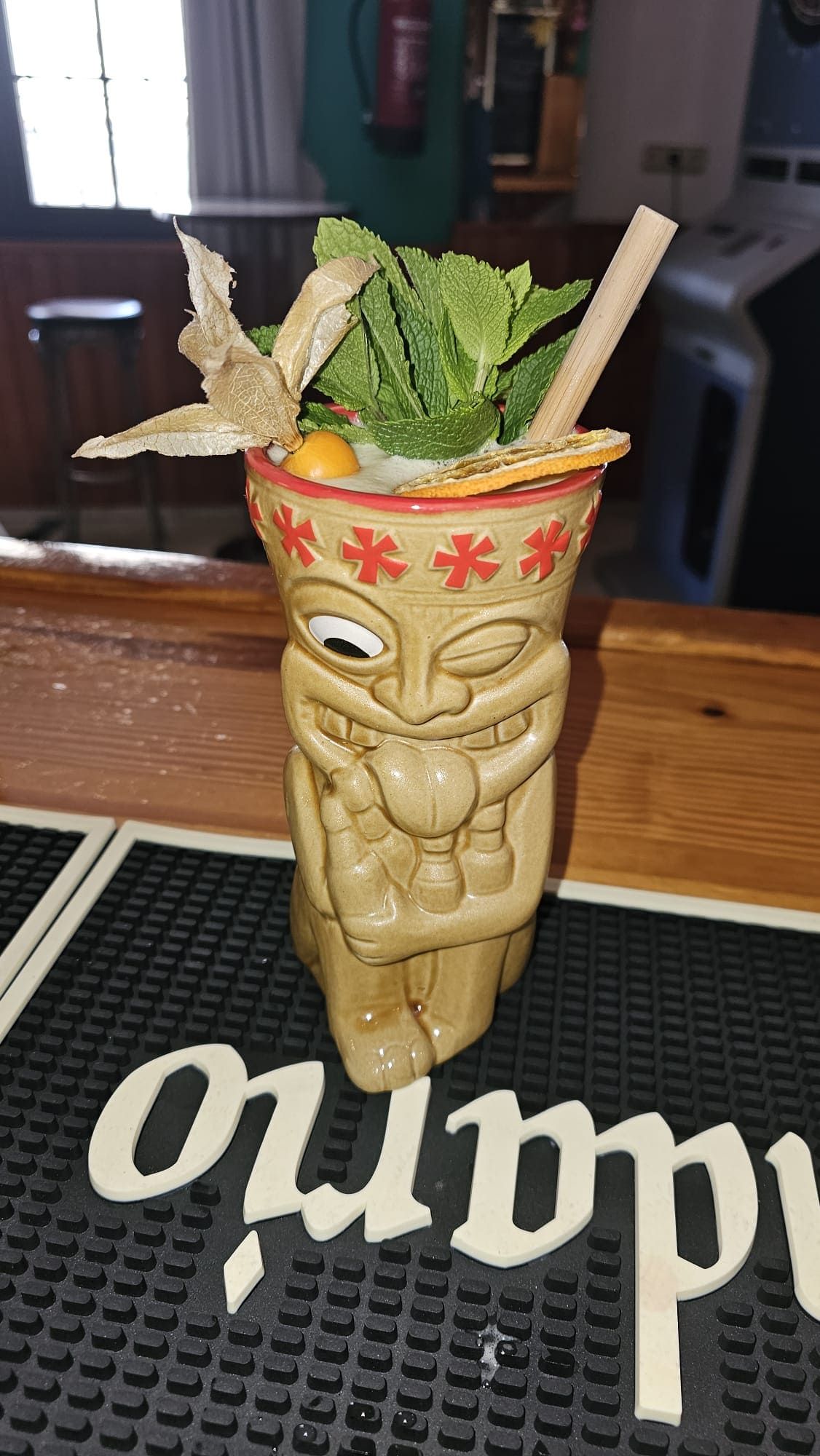 Una taza tiki con forma de personaje guiñando un ojo y sacando la lengua, adornada con menta fresca, una flor de physalis y una pajita sobre un mantel individual de bar.