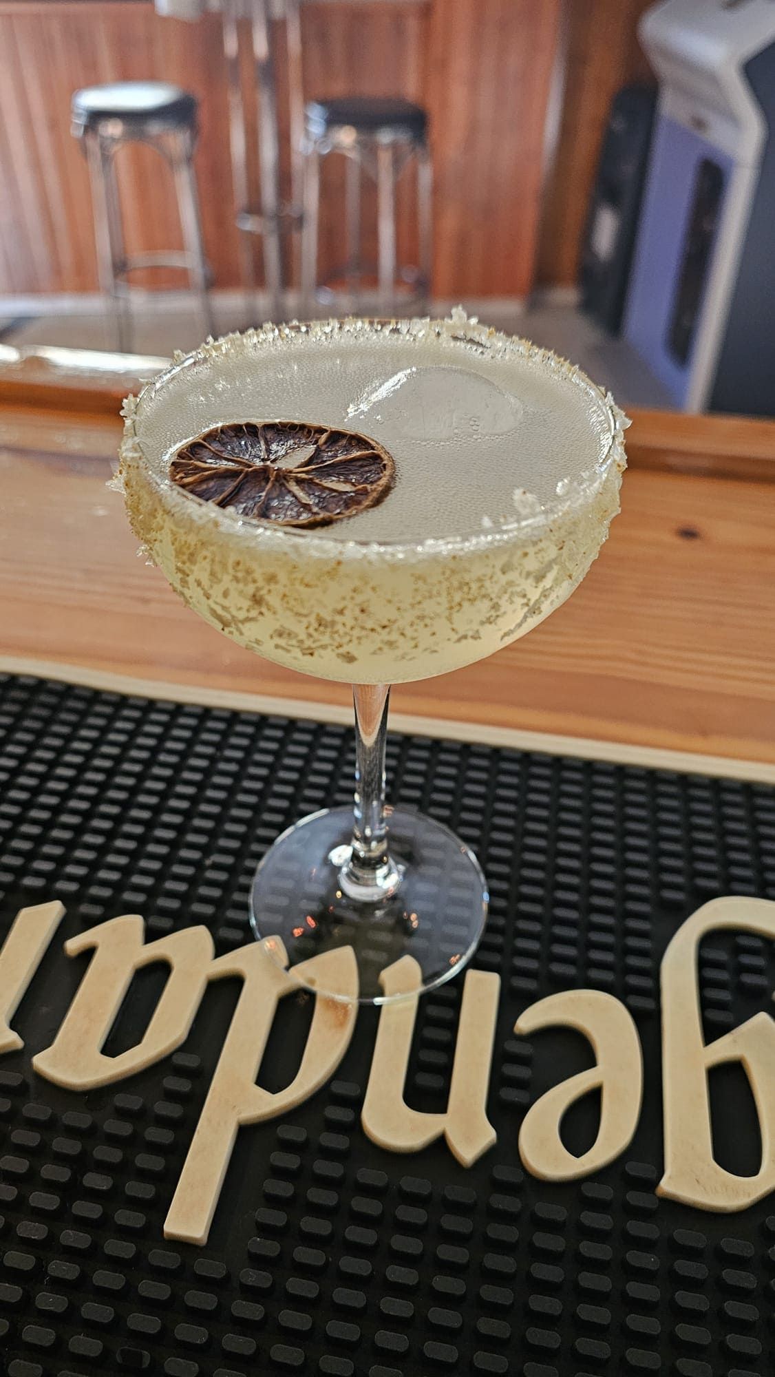 Un cóctel margarita servido en una copa tipo coupe con el borde escarchado con sal, adornado con una rodaja de lima deshidratada, sobre un mantel individual de bar.