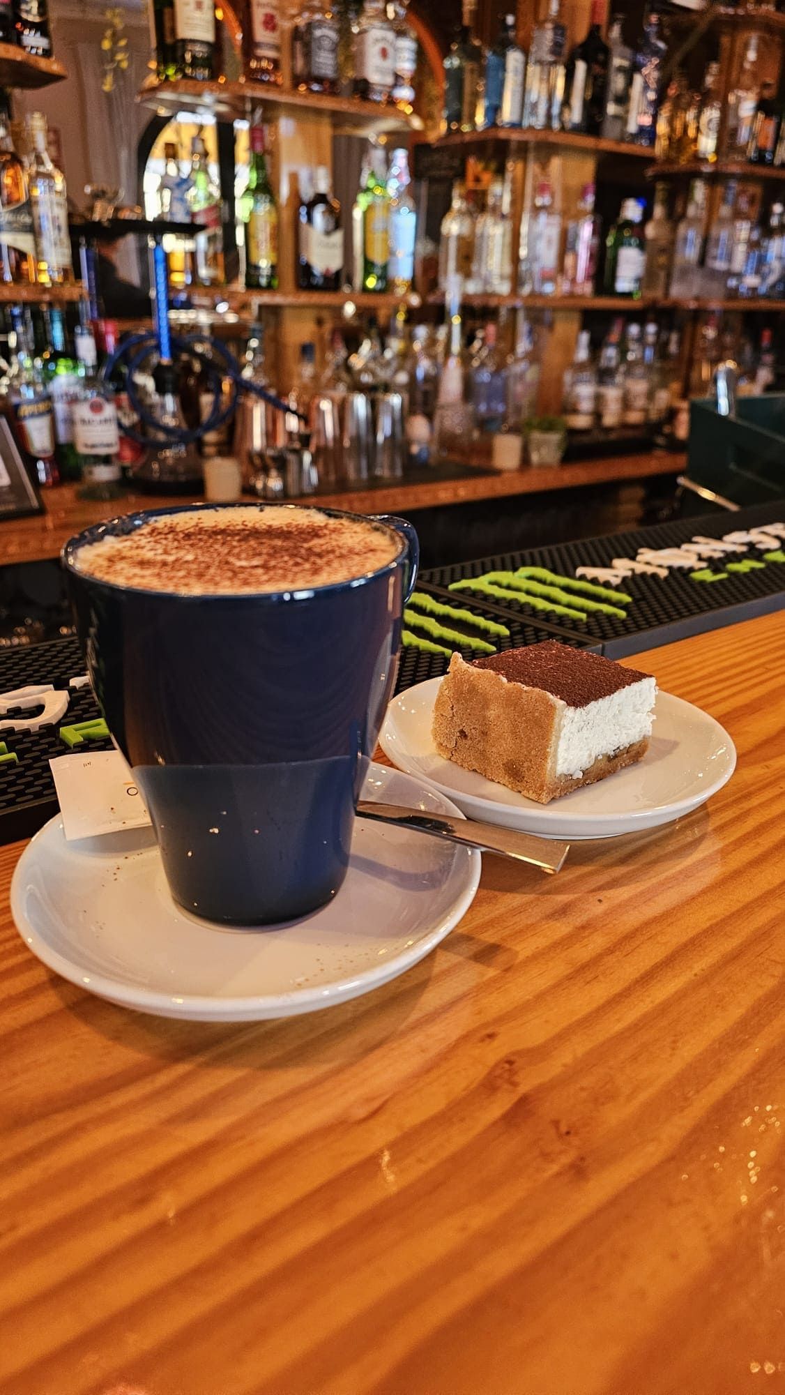Una taza azul de café y una pequeña porción de pastel reposan sobre platos en una barra de madera, frente a estantes repletos de licores.