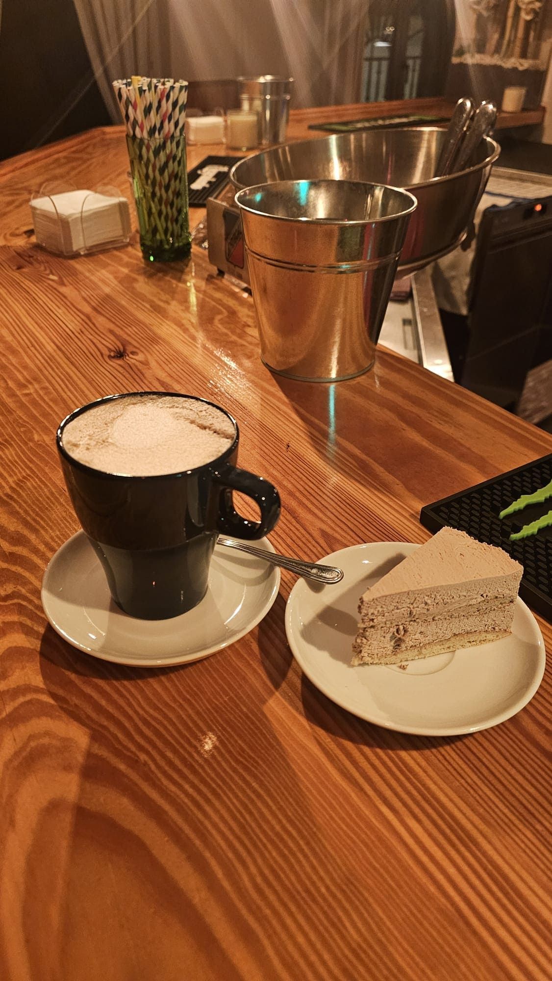 Una taza de café con espuma y una porción de pastel sobre platos blancos reposan en una barra de madera, con recipientes de metal cerca.