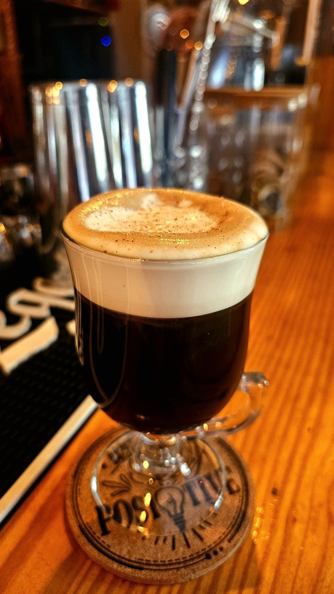 Un café irlandés con una espesa capa de crema, servido en un vaso con asa sobre un posavasos de corcho en una barra de madera.