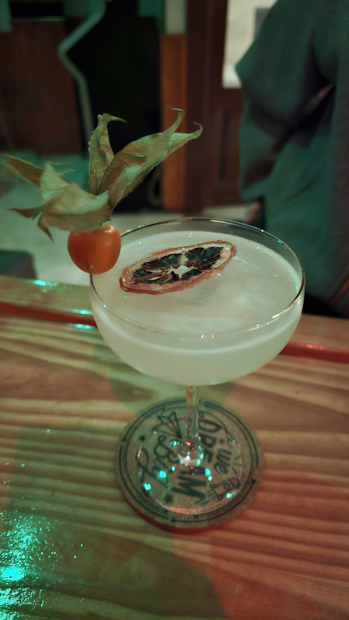 Un cóctel servido en una copa tipo coupe, adornado con una flor de physalis y una rodaja de cítricos secos, sobre una barra de madera.
