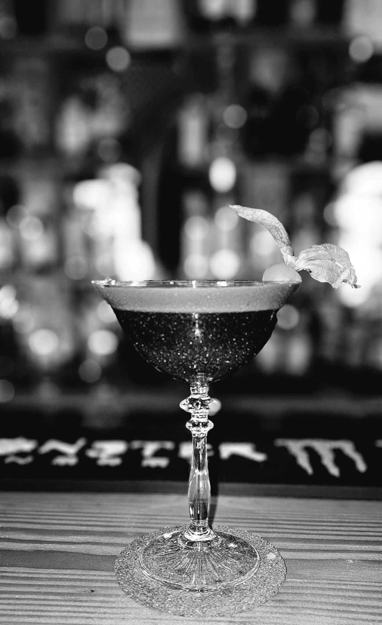 Una fotografía en blanco y negro de una elegante copa de cóctel con tallo, adornada con una guarnición, sobre la barra de un bar.