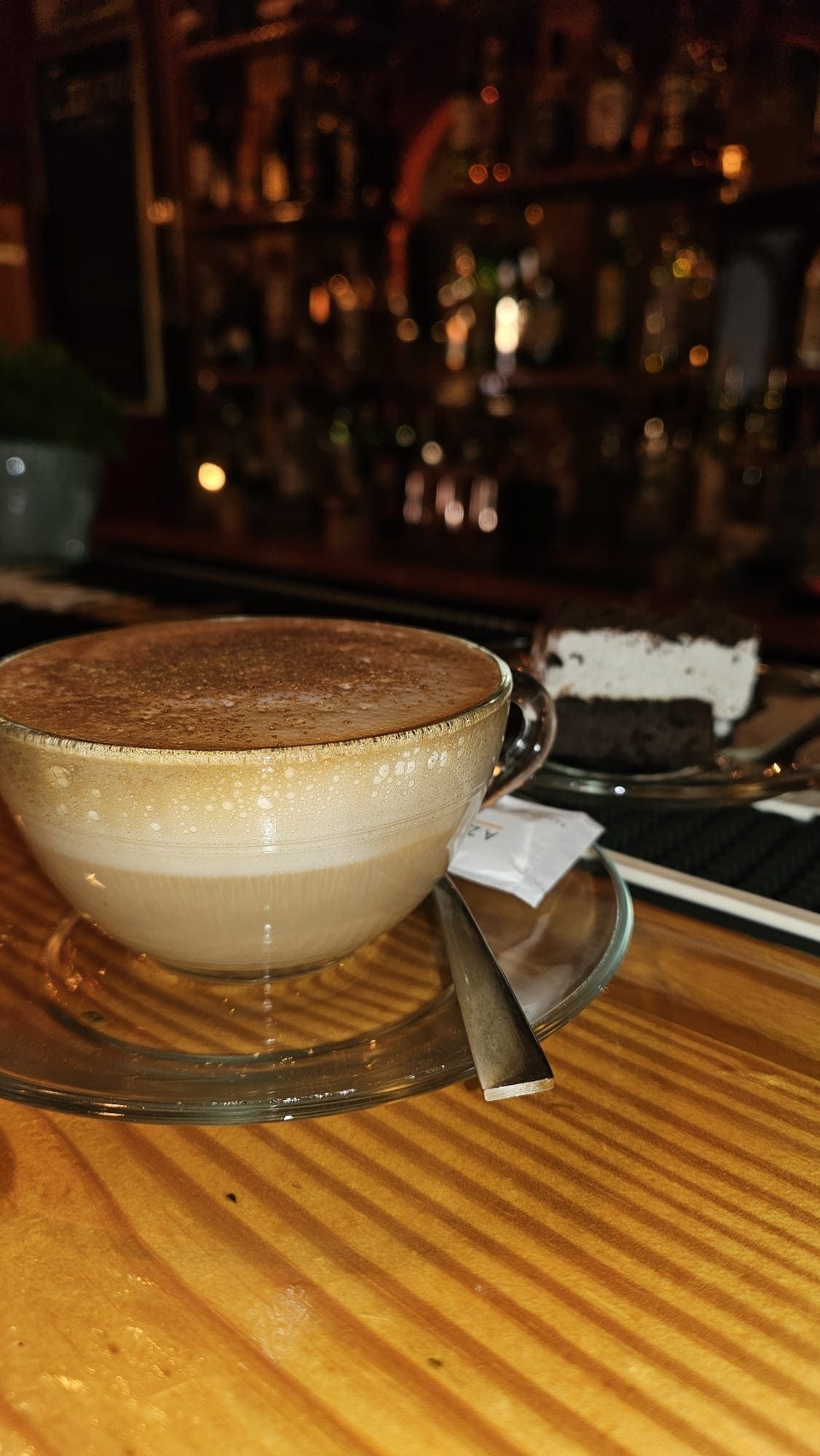 Primer plano de un capuchino en una taza de cristal sobre una mesa de madera, junto a una pequeña porción de pastel en un bar con iluminación tenue.