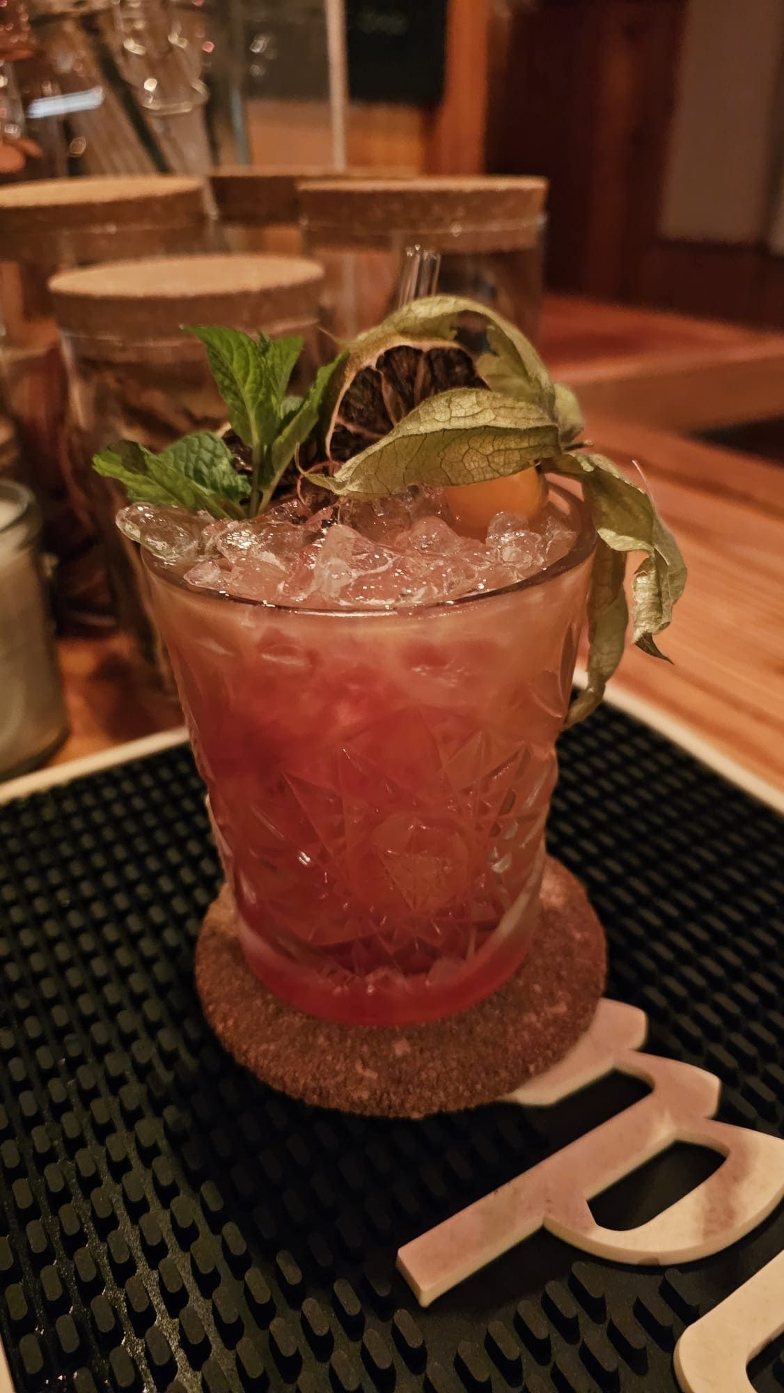 Un cóctel adornado con menta y una baya dorada en un vaso texturizado sobre un posavasos de corcho encima de una alfombrilla de goma para bar.