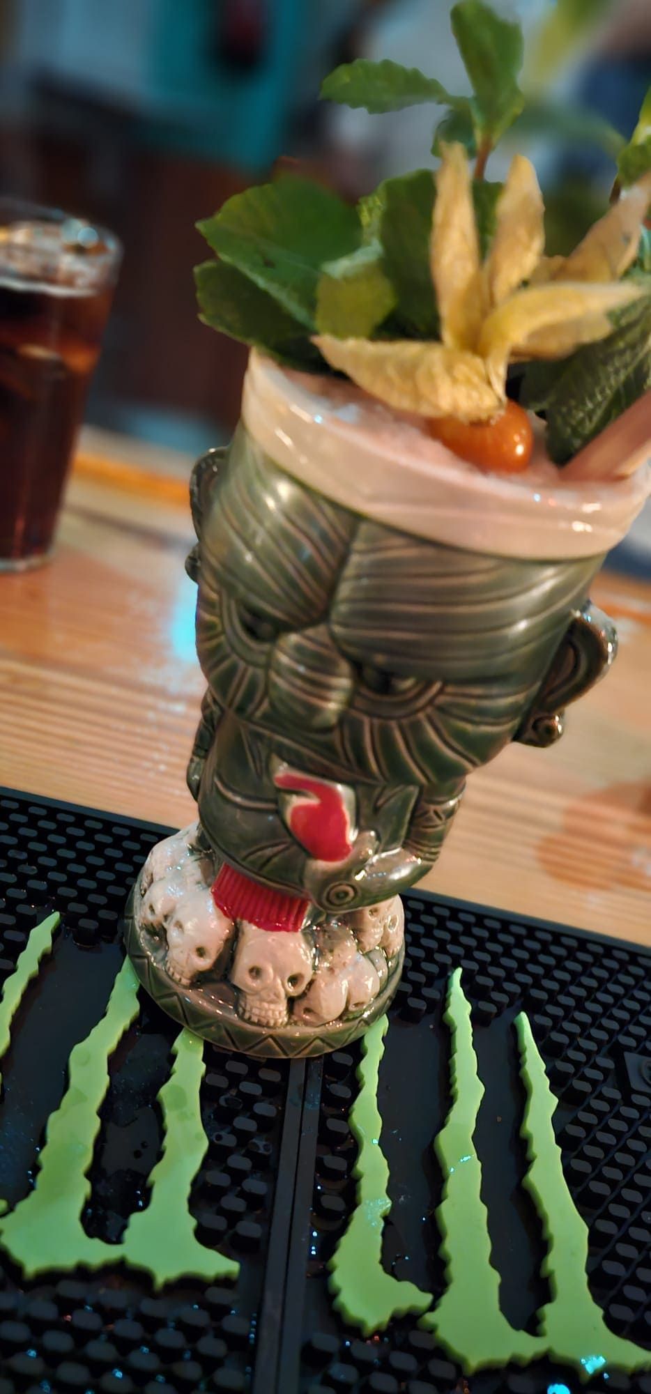 Una detallada taza tiki llena de una bebida tropical, adornada con menta y piña, descansa sobre una esterilla de Monster Energy.