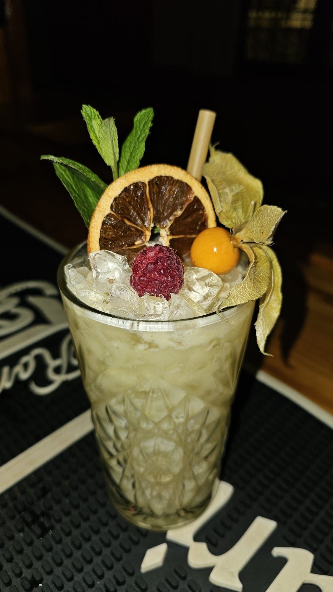 Un cóctel refrescante en una copa decorada con motivos florales, adornado con menta, una rodaja de naranja deshidratada, una frambuesa y una baya dorada.