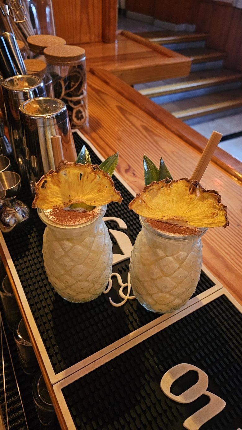 Dos vasos con forma de piña, llenos de cremosos cócteles tropicales, cada uno adornado con una rodaja de piña, sobre un mantel individual de bar.