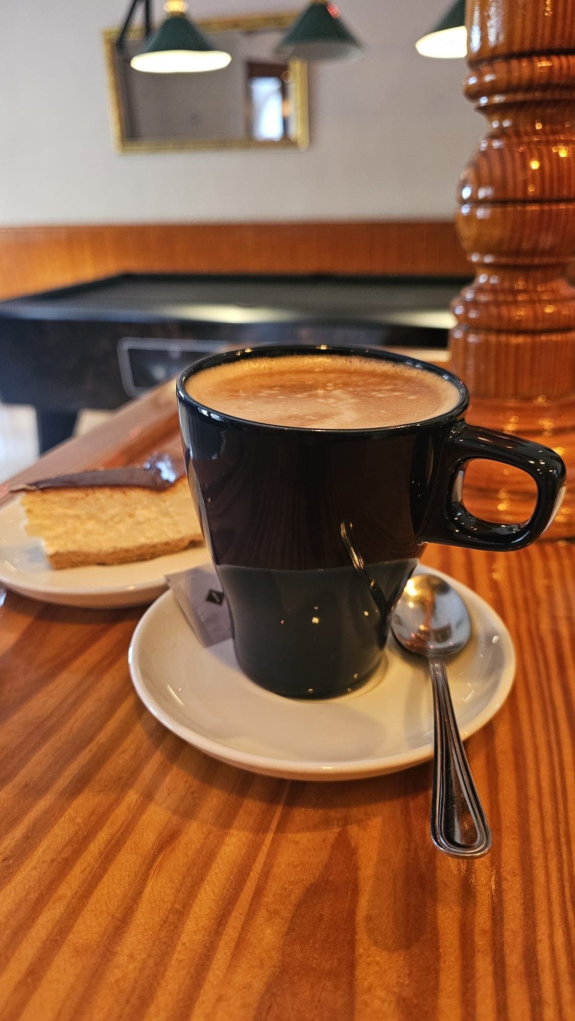 Una taza de café negra sobre un platillo blanco con una cuchara y una porción de pastel sobre una barra de madera en una sala de billar.