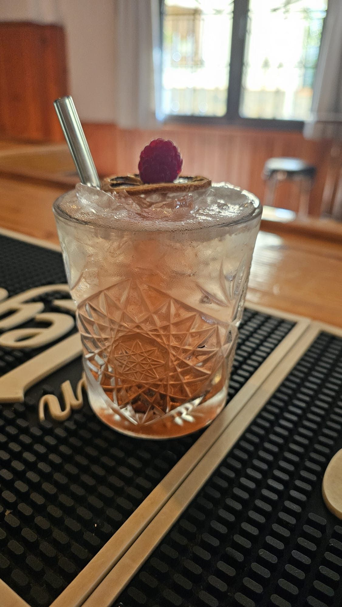 Un cóctel en un vaso estampado, adornado con una rodaja de cítricos deshidratados y una frambuesa fresca, reposa sobre un mantel individual de bar.