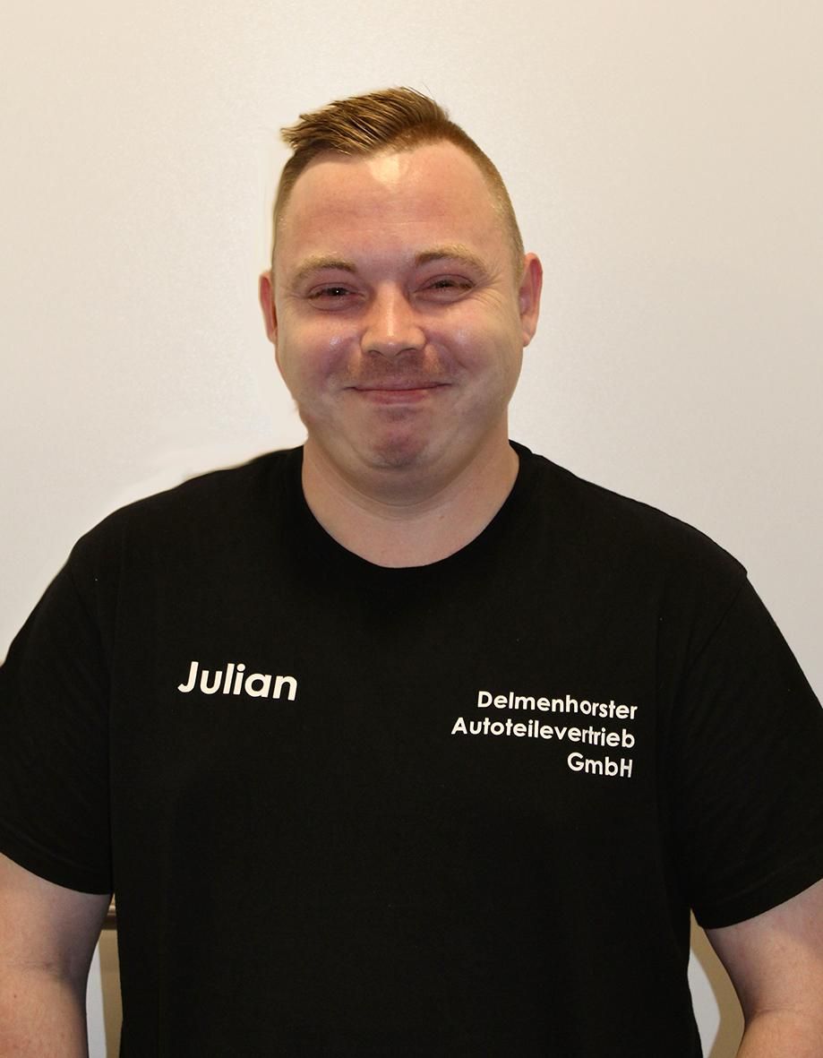 Eine Person lächelt in die Kamera und trägt ein schwarzes T-Shirt mit der Aufschrift „Julian“ und dem Firmennamen auf der Vorderseite.