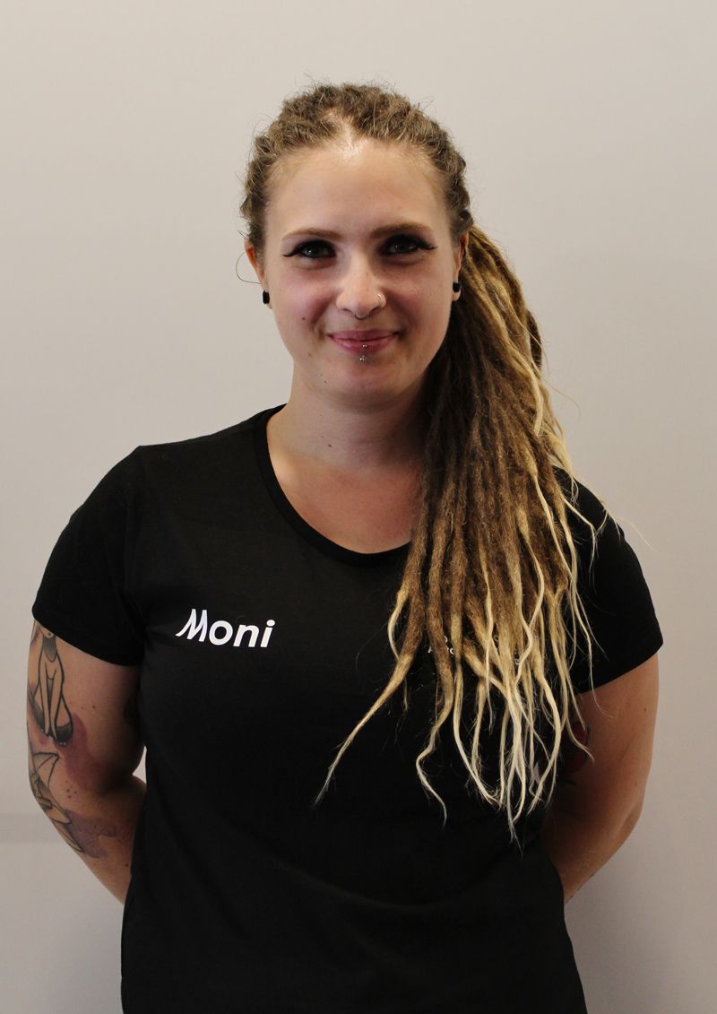 Frau mit Dreadlocks, trägt ein schwarzes T-Shirt mit der Aufschrift „Moni“ und steht vor einer grauen Wand.