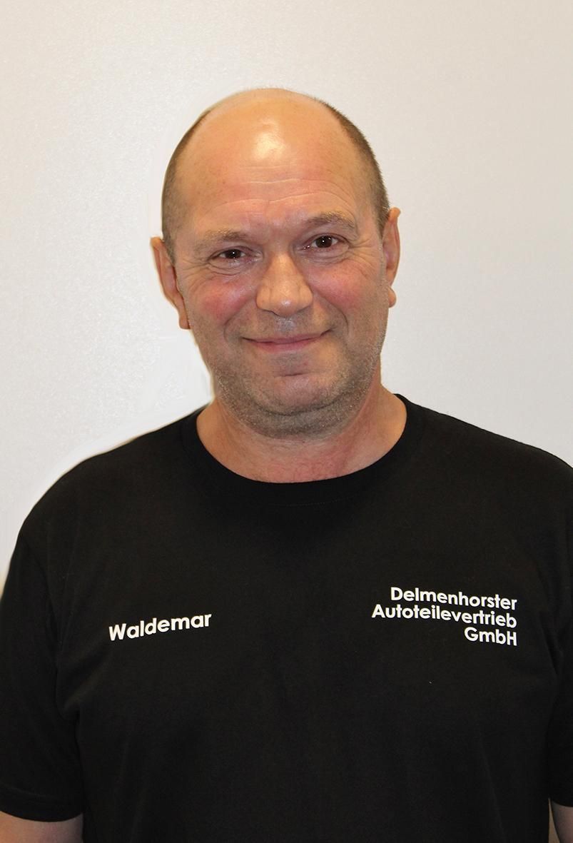 Eine Person in einem schwarzen T-Shirt mit Firmenlogo lächelt vor einem schlichten weißen Hintergrund.