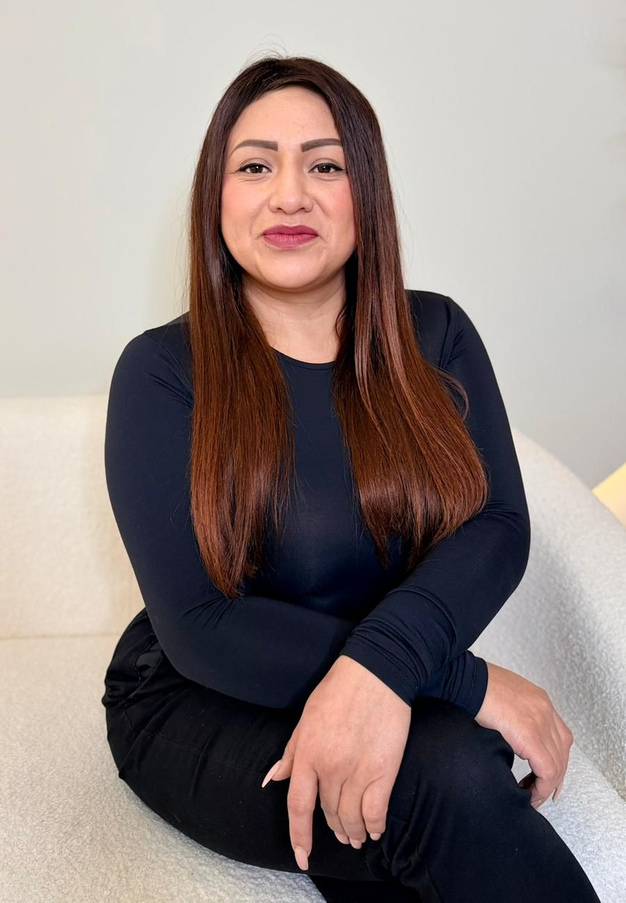 Mujer con cabello largo y castaño, vestida con una camisa negra de manga larga, sentada en un sofá blanco, sonriendo.