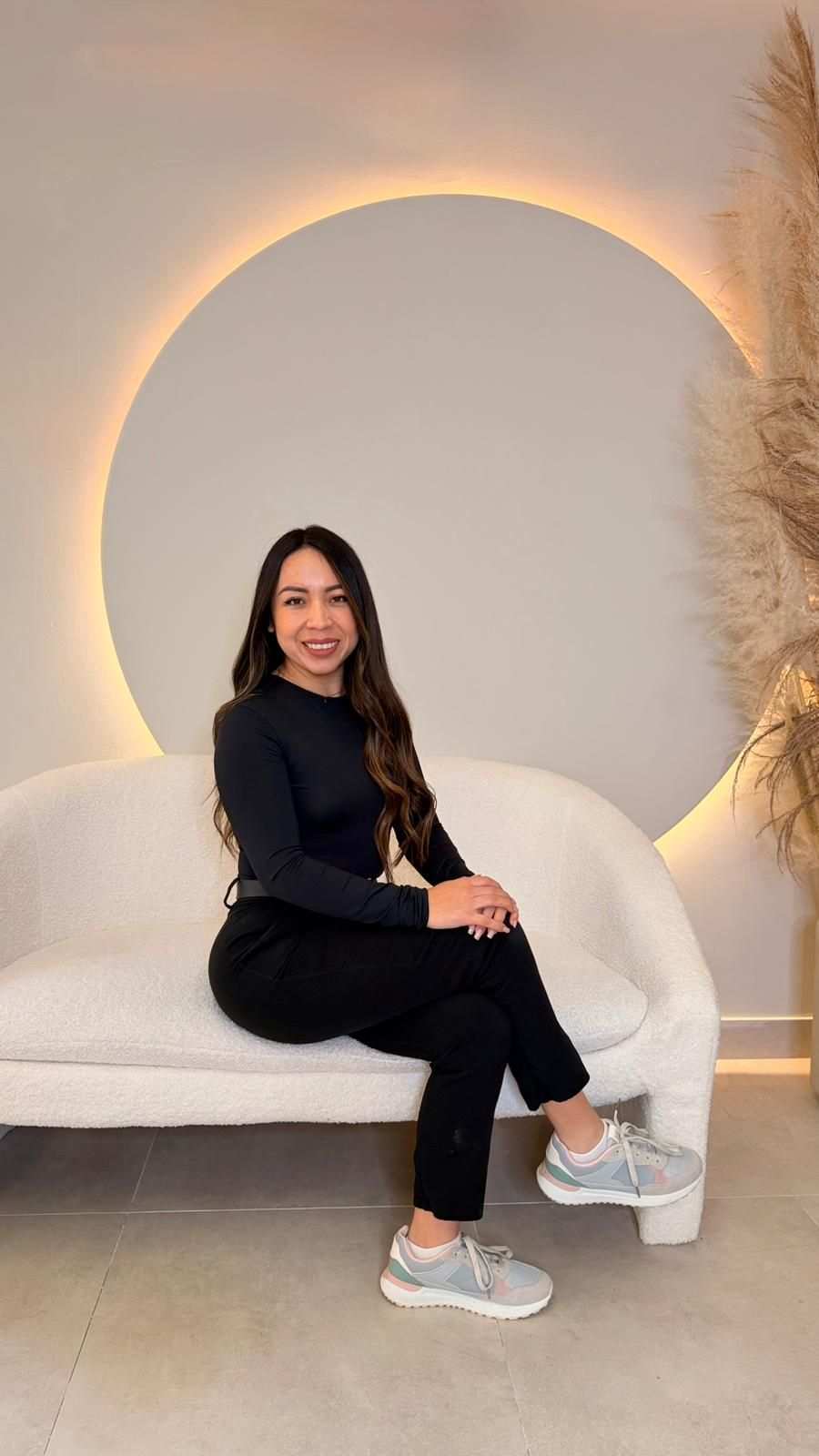 Una mujer vestida de negro se sienta en un sofá curvo blanco, sonriendo frente a una luz circular en una pared blanca.