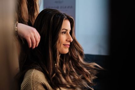 Mujer con cabello largo y ondulado sonriendo de perfil en interiores, con una mano borrosa en primer plano.