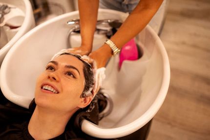 Una mujer se está lavando el pelo en el lavabo de una peluquería, sonriendo mientras mira las manos de la estilista.
