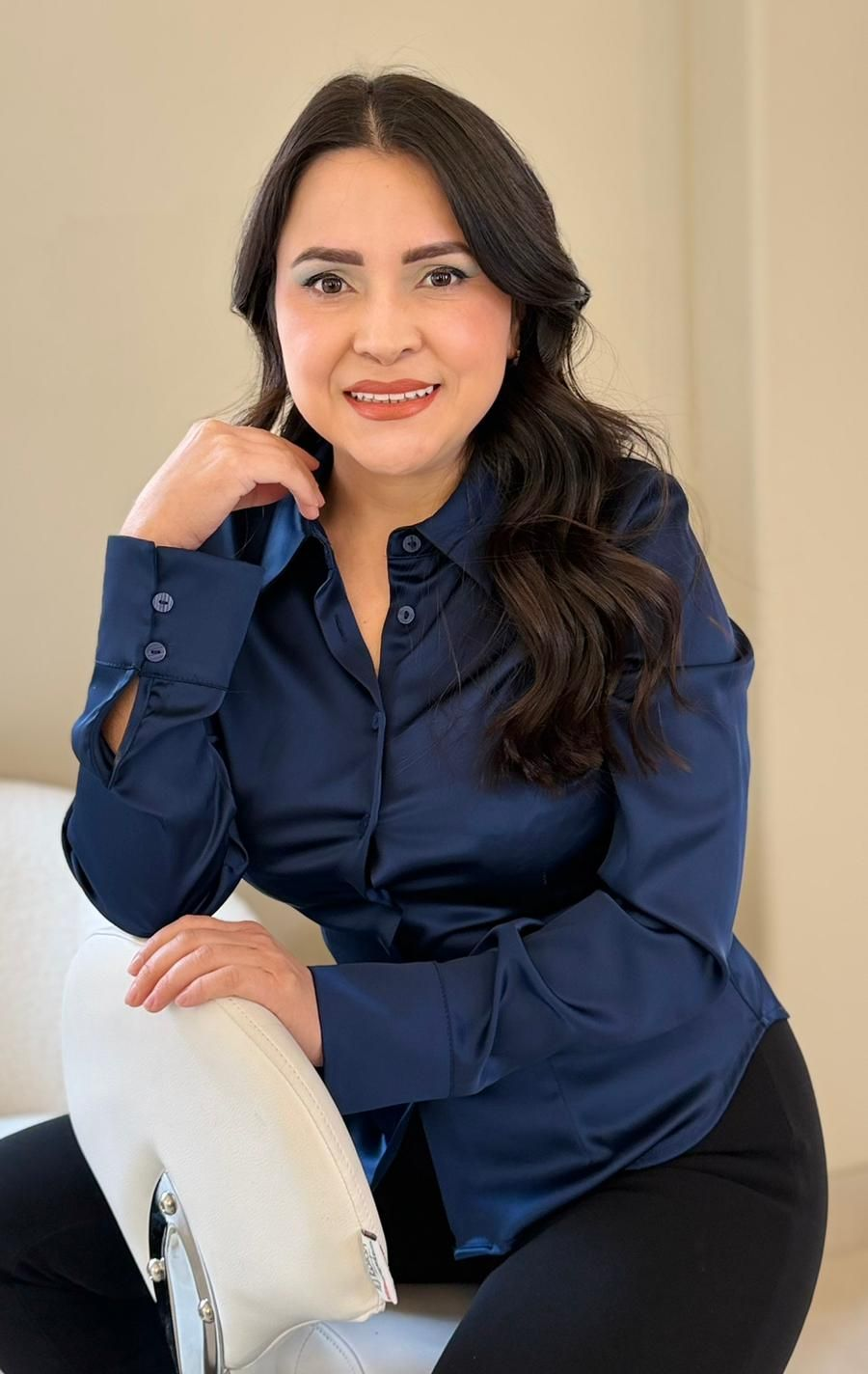 Mujer con camisa azul marino y pantalón negro, sonriente, sentada en una silla blanca.