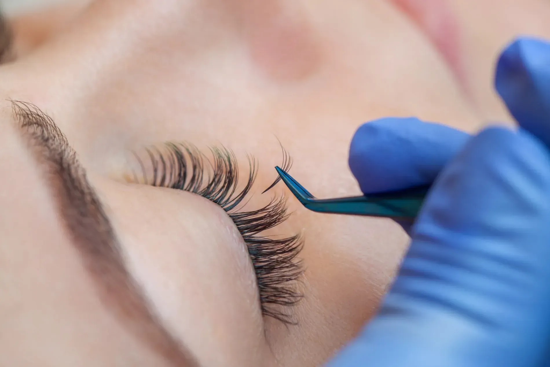 Extensión de pestañas aplicada con pinzas mediante una mano enguantada sobre un ojo cerrado.