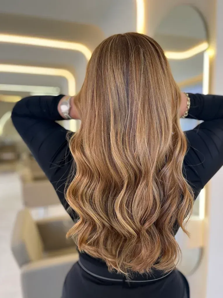 Mujer con cabello castaño largo, ondulado y con reflejos. Manos sujetando el cabello; vestida de negro. Salón luminoso.