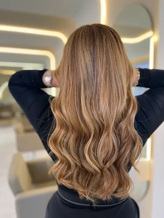 Mujer con cabello castaño largo y ondulado de pie con las manos detrás de la cabeza en el interior de un salón de belleza moderno.