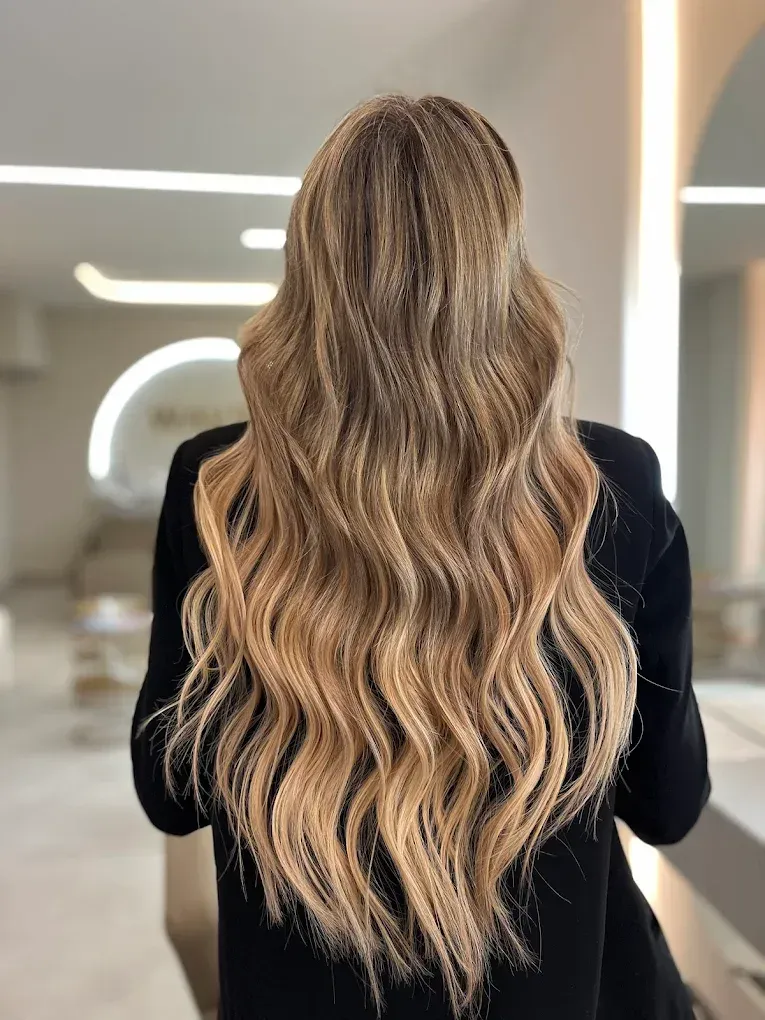 Mujer con cabello rubio largo, ondulado y rizado, vestida con un top negro, en un interior lleno de luz.