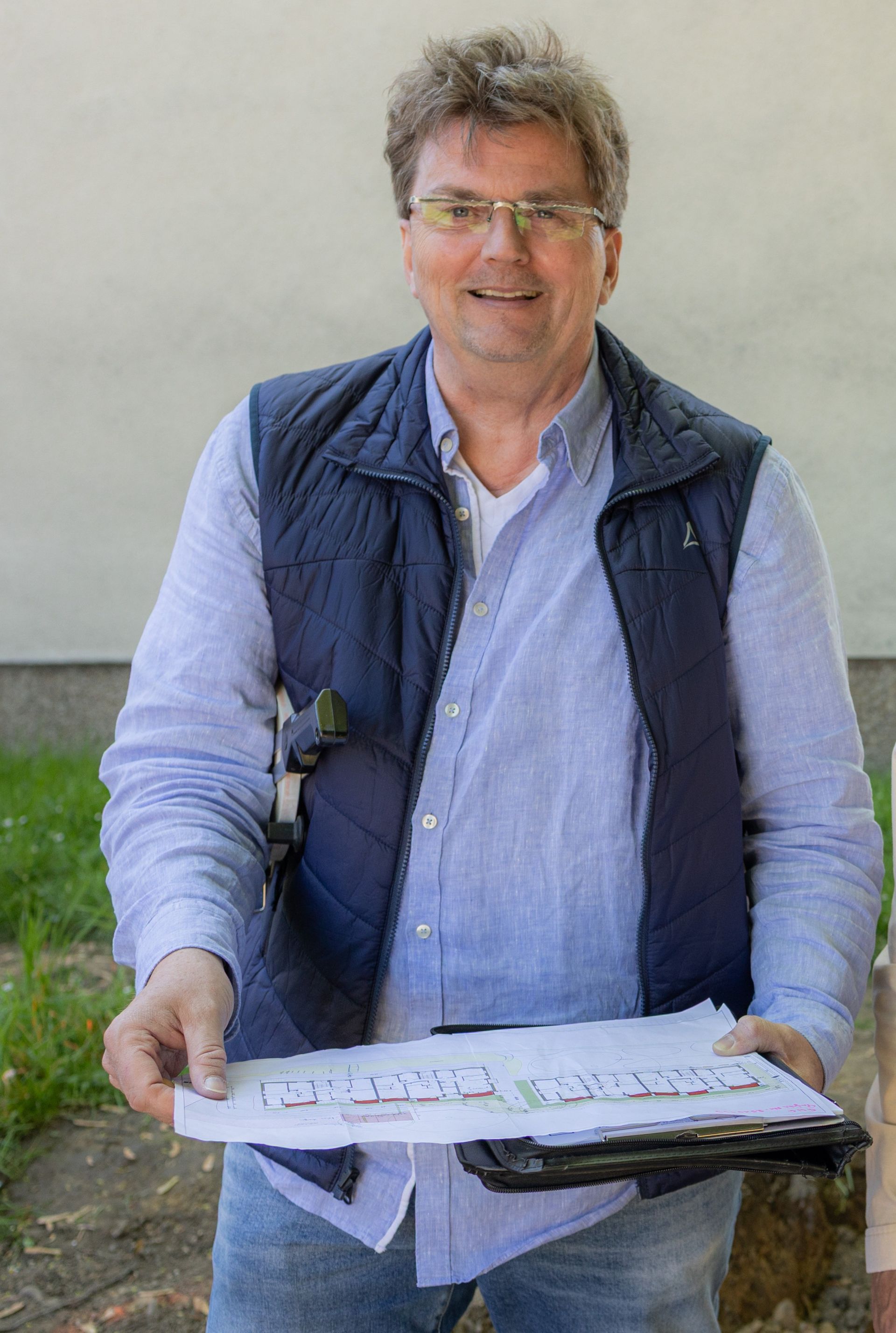 Ein Mann mit Brille, der Papiere in der Hand hält und Weste und zugeknöpftes Hemd trägt, lächelt im Freien.
