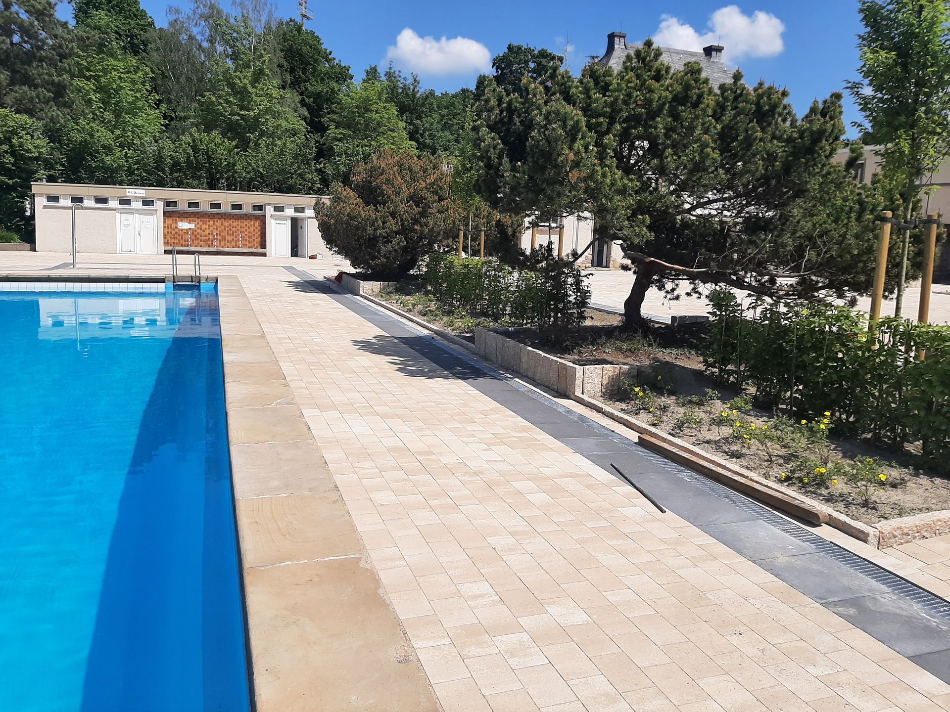 Am Pool mit blauem Wasser, beigem Gehweg und Umkleidekabinen. Im Hintergrund sind Bäume und ein Gebäude zu sehen.