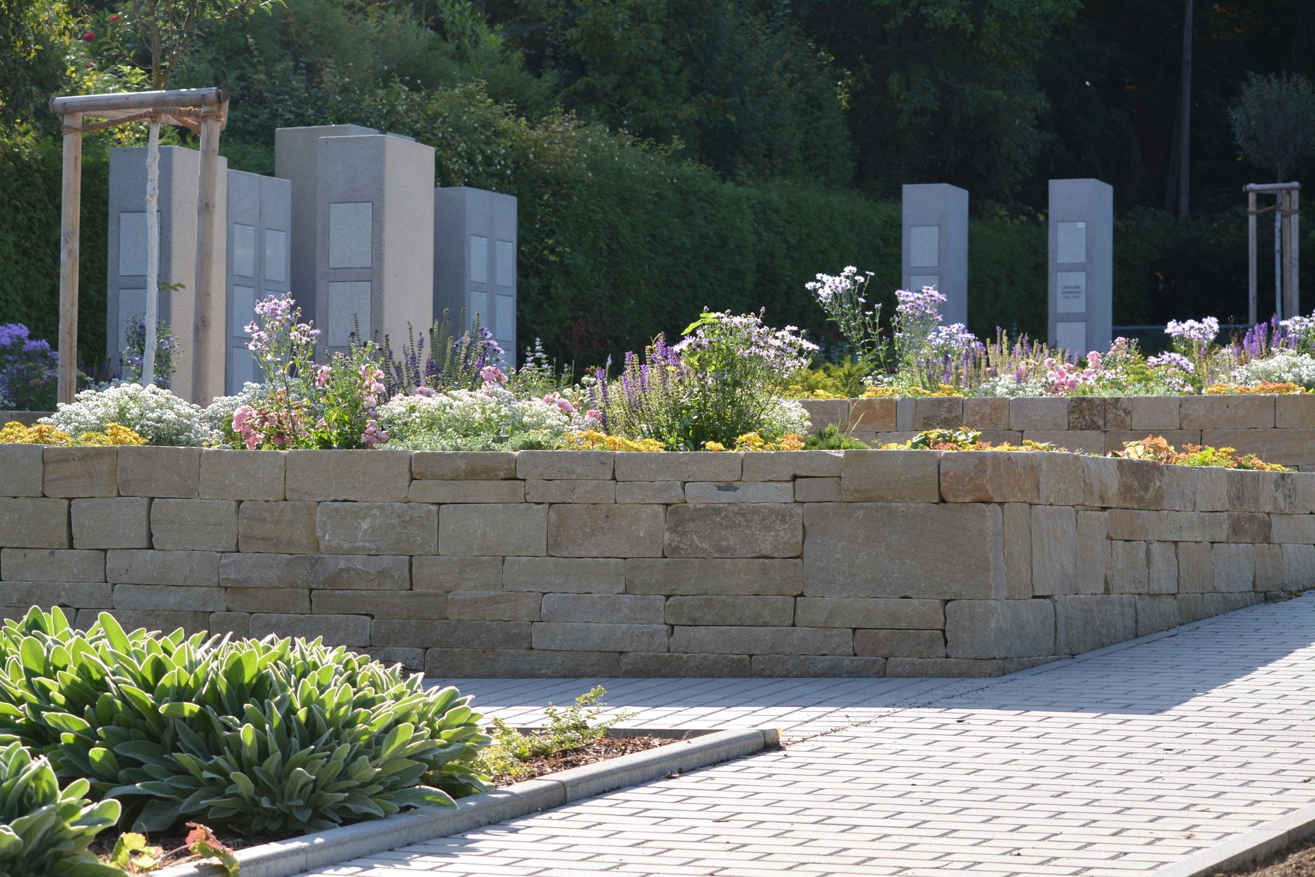 Steinmauern mit Blumenbeeten vor Gedenksäulen.