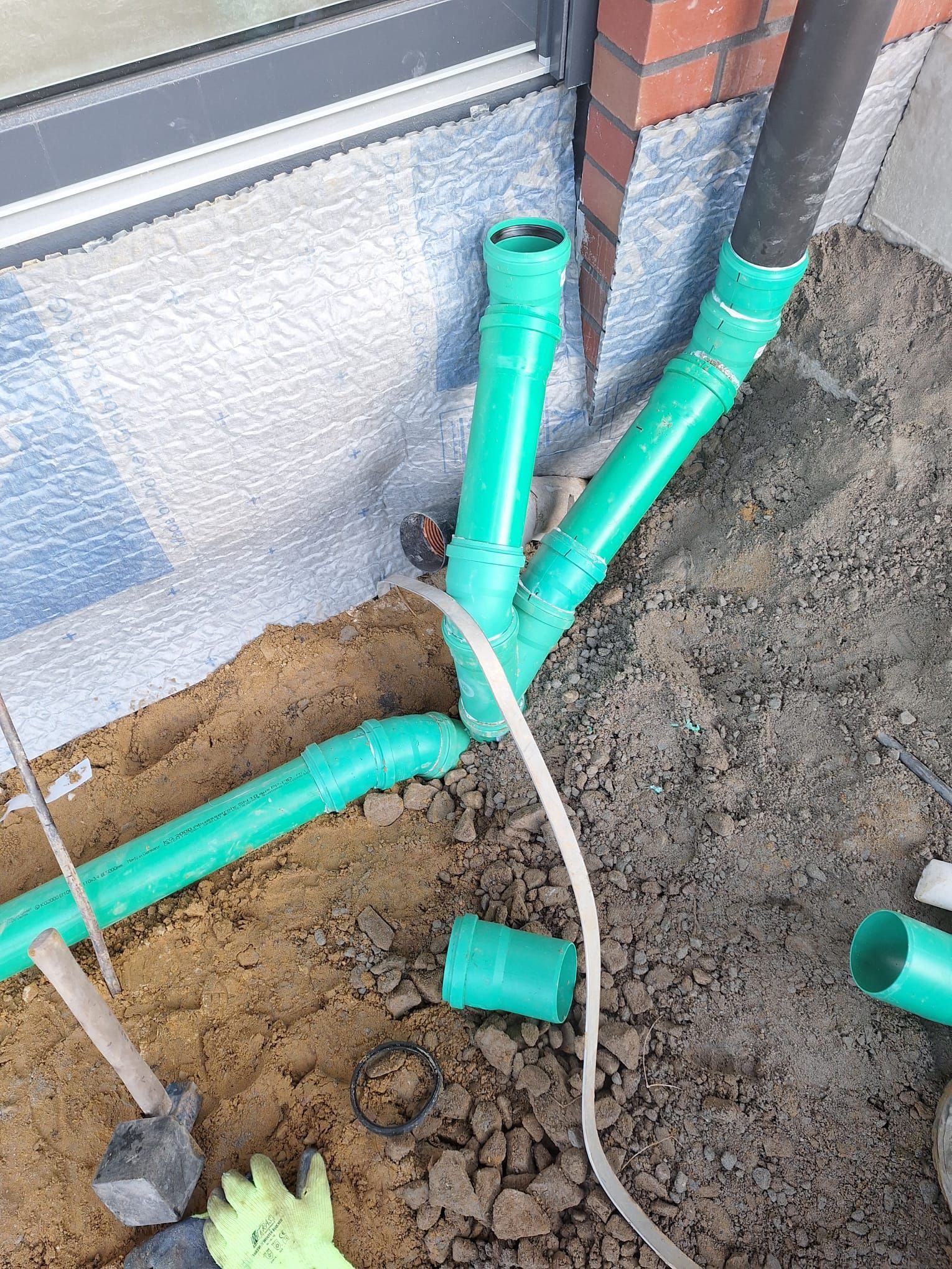 Grüne Kunststoff-Abflussrohre sind in der Nähe des Fundaments eines Gebäudes miteinander verbunden; im Erdreich sind Bauwerkzeuge sichtbar.