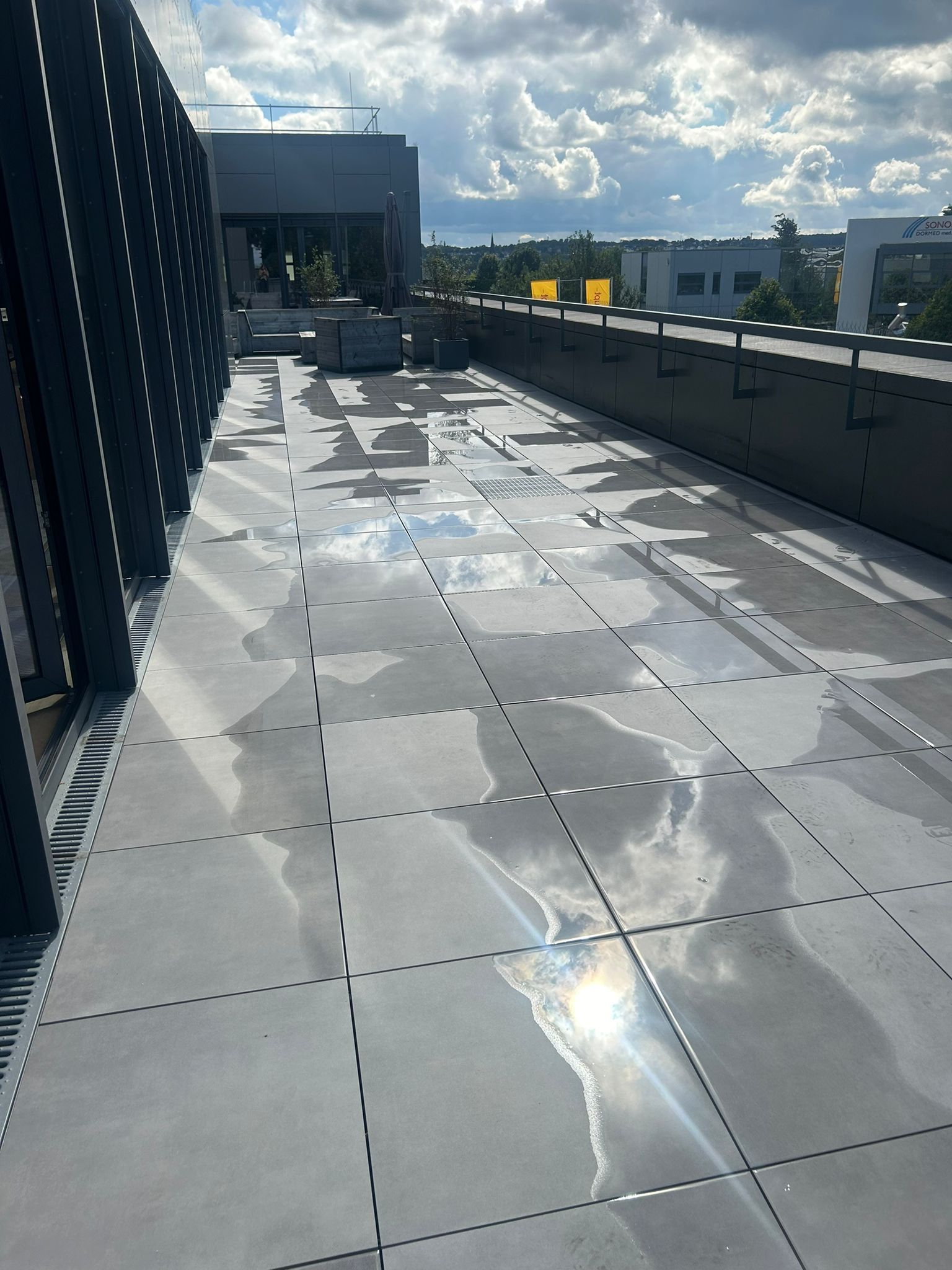 Eine feuchte Dachterrasse mit grauem Fliesenboden, die Sonnenlicht und einen bewölkten Himmel reflektiert.