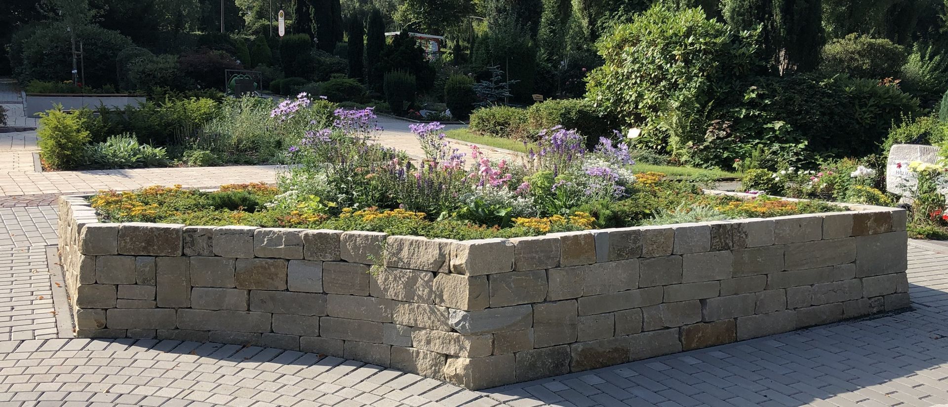 Niedrige Steinmauer mit farbenfrohen Blumen und Grünpflanzen. Im Hintergrund gepflasterter Weg und Bäume.