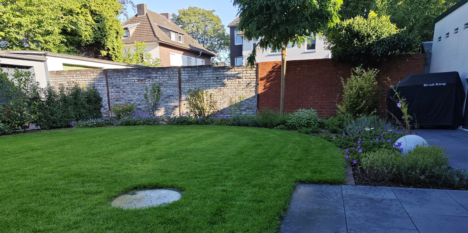 Grüner Rasen mit einer Backsteinmauer und Bäumen im Hintergrund. Rechts befindet sich eine gepflasterte Terrasse.
