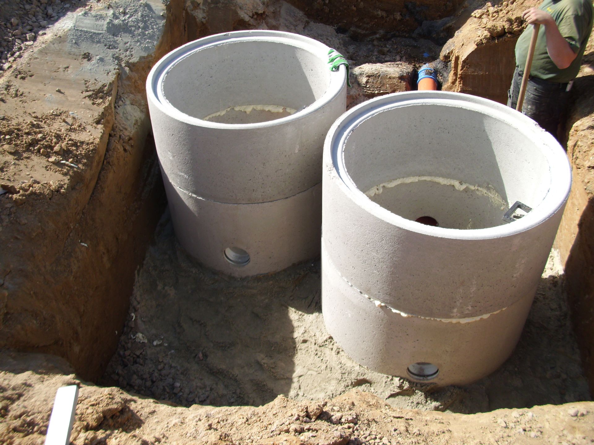 Zwei Betonringe in einem Erdgraben, vermutlich für eine Klärgrube. Eine Person steht daneben.