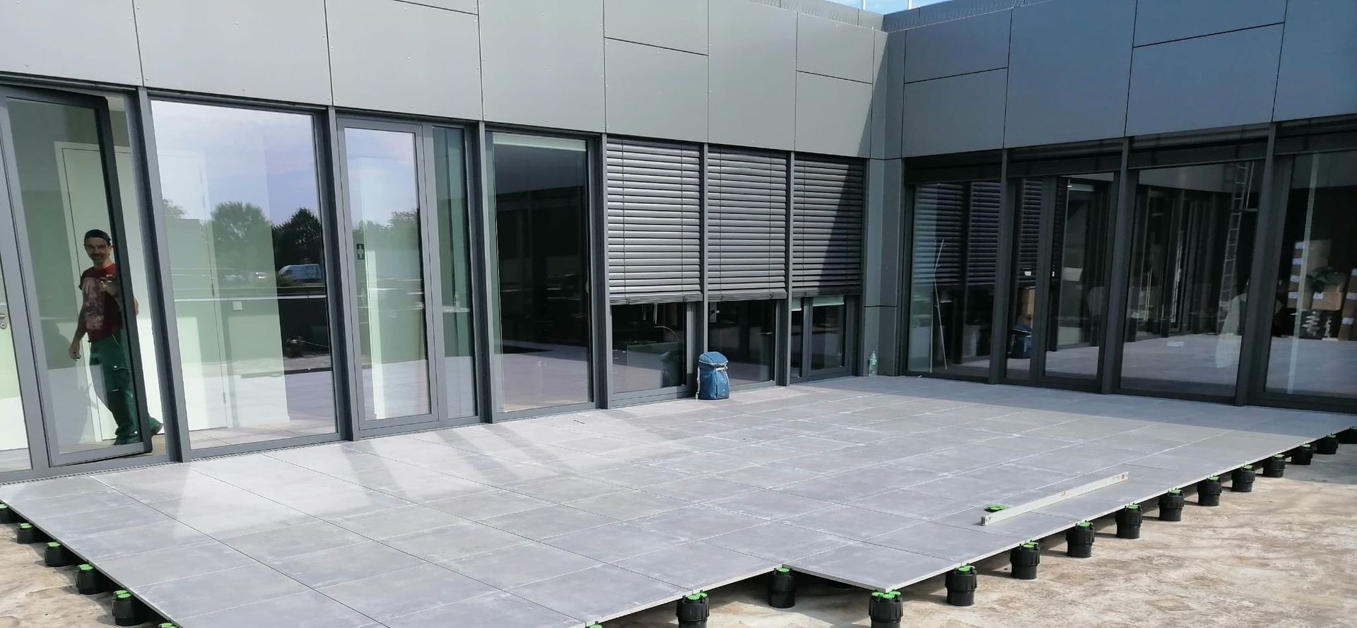 Außenansicht eines Gebäudes mit Glastüren, auf dem graue Verbunddielen verlegt werden. Eine Person arbeitet im Inneren.
