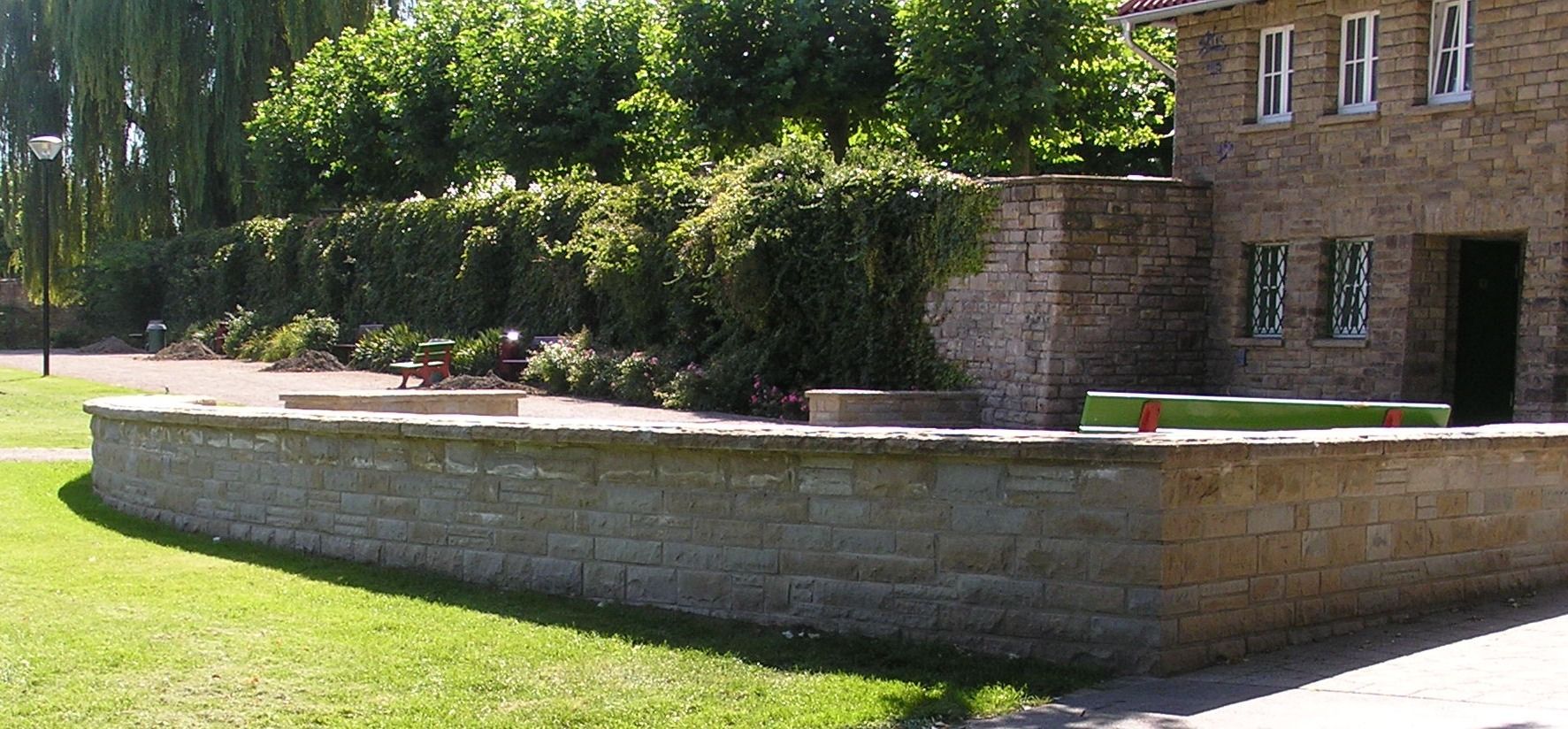 Steinmauer und Gebäude, umgeben von Grün, sonniger Tag.