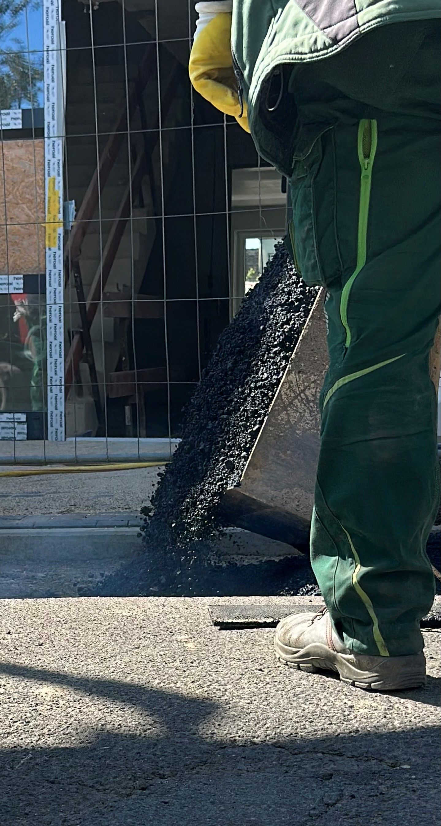 Eine Person in grüner Arbeitskleidung gießt Asphalt auf eine Fläche in der Nähe eines Gebäudes mit Fenstern.