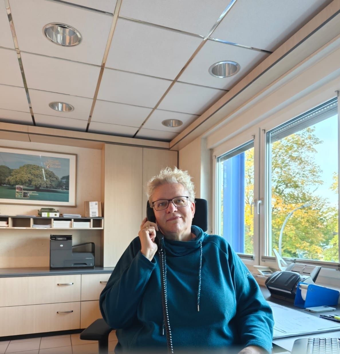 Eine Frau telefoniert an einem Schreibtisch in einem Büro mit Fenster und einem Gemälde an der Wand.